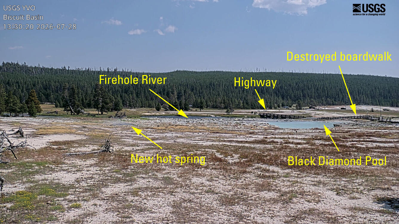 remote camera view of barren ground with a blue hot spring in the middle distance and a forested ridge in the background