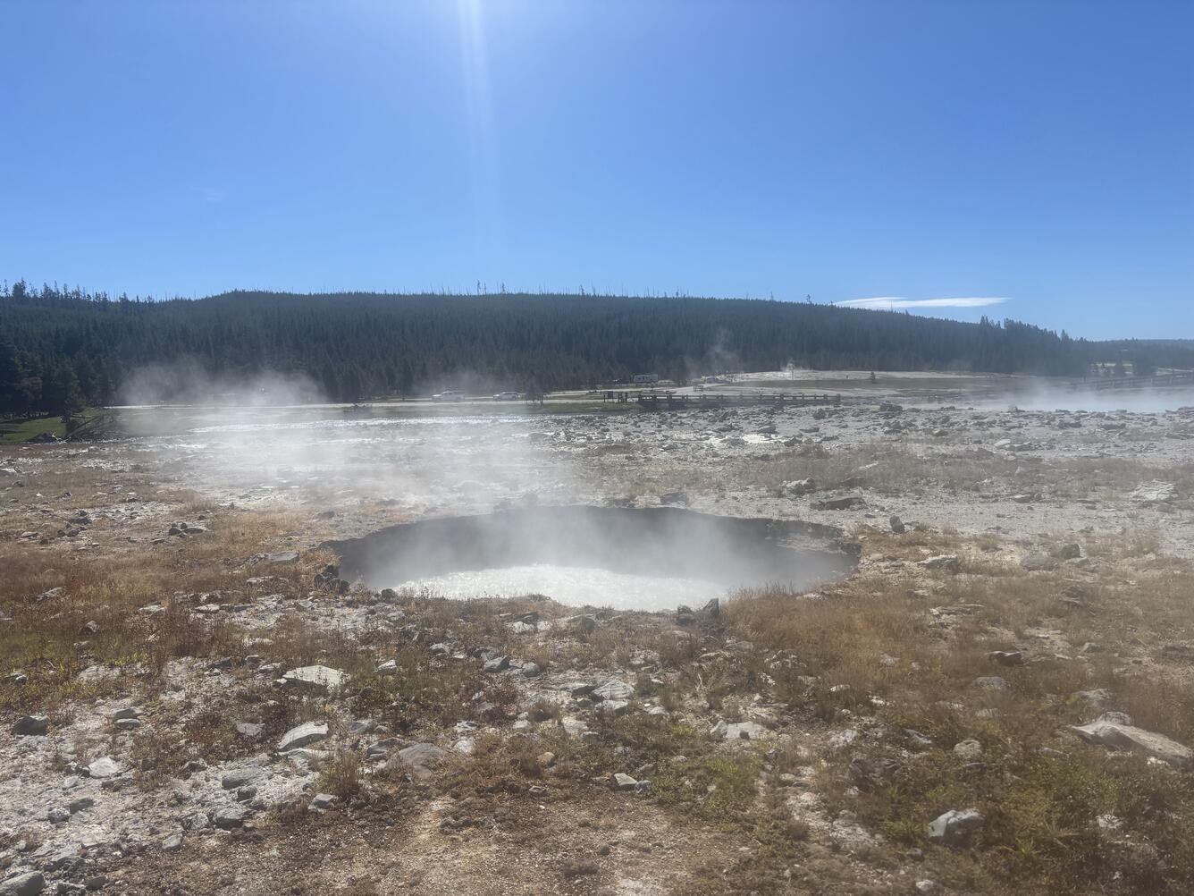 Circular pool of boiling water in a barren area strewn with rocks with a forested ridge in the distance under blue sky.