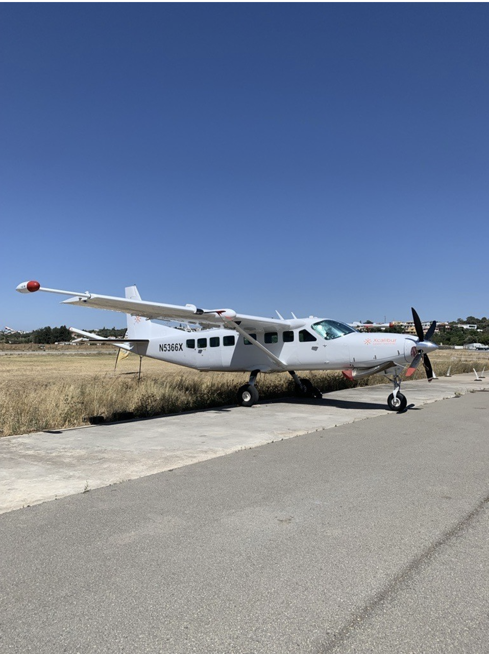 FIxed wing aircraft with boom extension, parked on airstrip with tall grass behind.