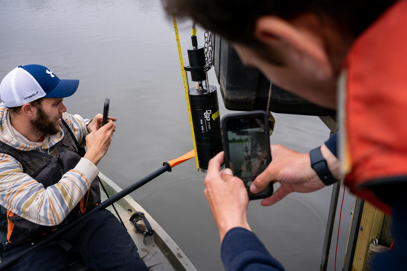 Two people photograph a scientific instrument over the water with their phones.