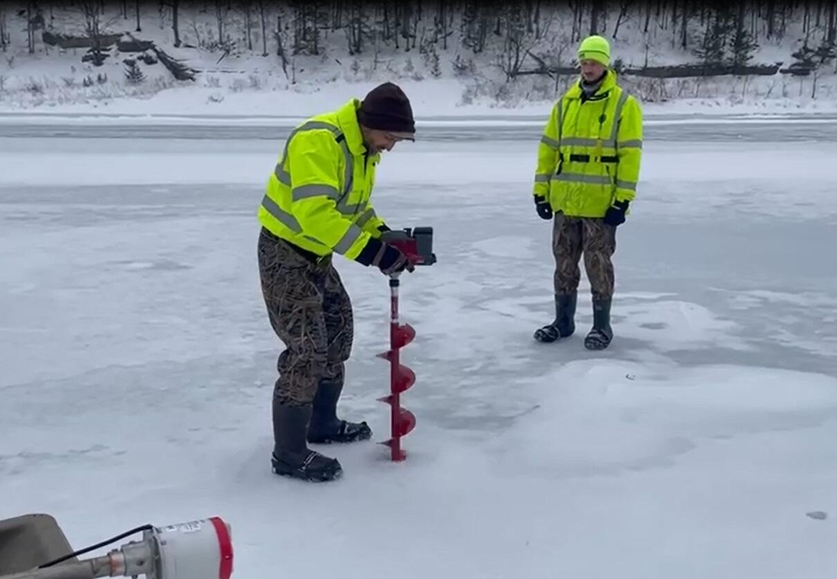 Two scientists standing on a frozen river drilling a hole to check the flow below