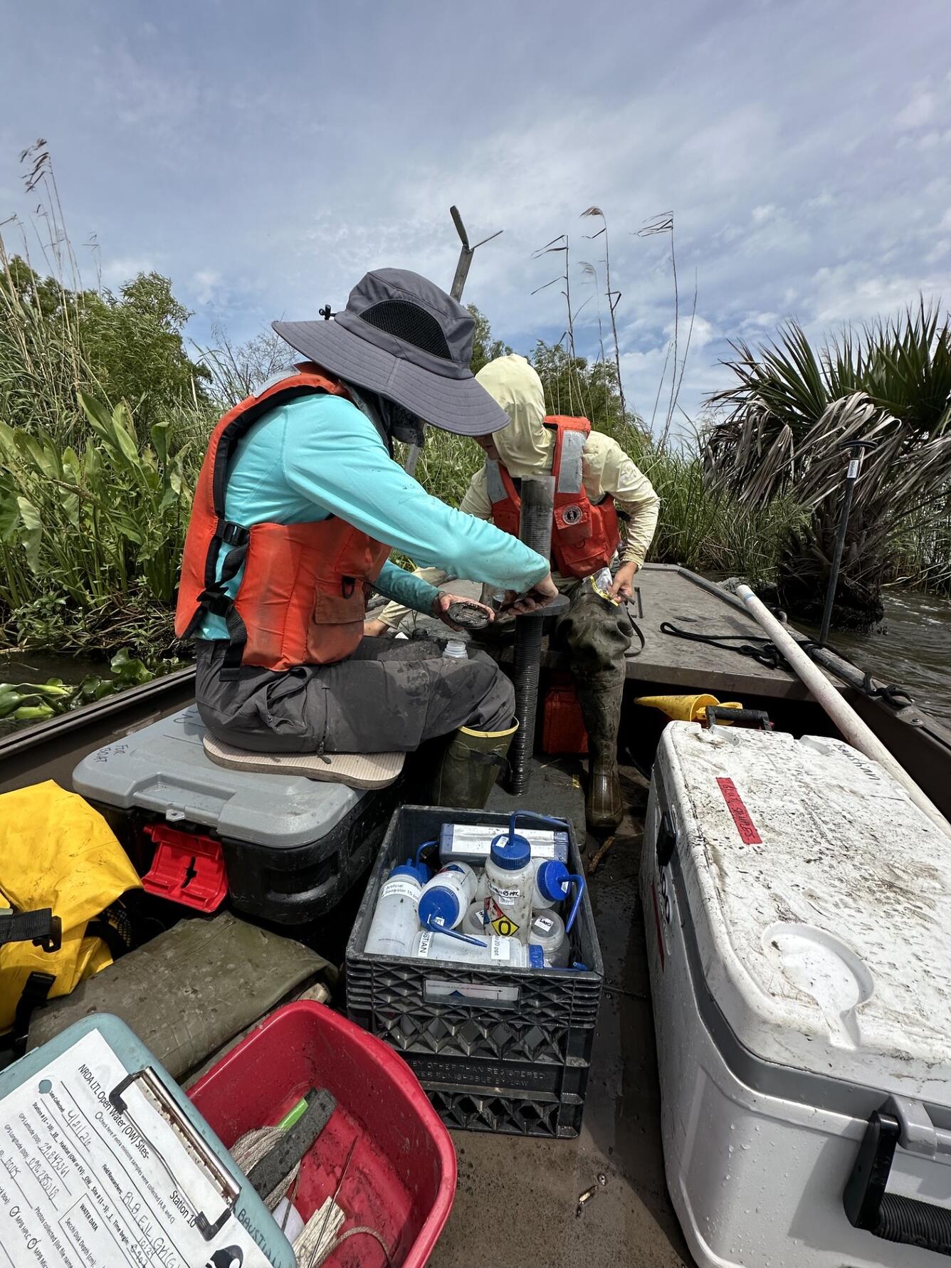 Two scientists sit in a boat surrounded by coolers and equipment. They both look down at their hands as they take samples from sediment cores collected at the marsh edge. Plants can be seen in the background.
