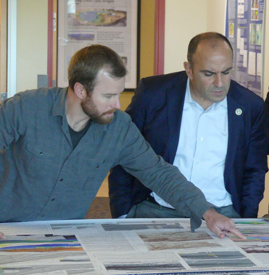 A man points out something on a map, to another man who looks on.