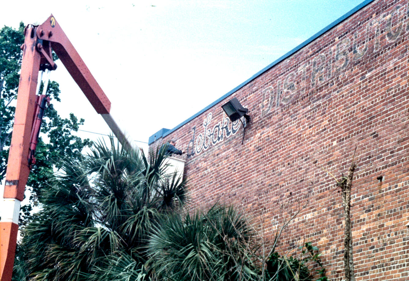 A worker on a lift paints over faded text on the side of a brick building