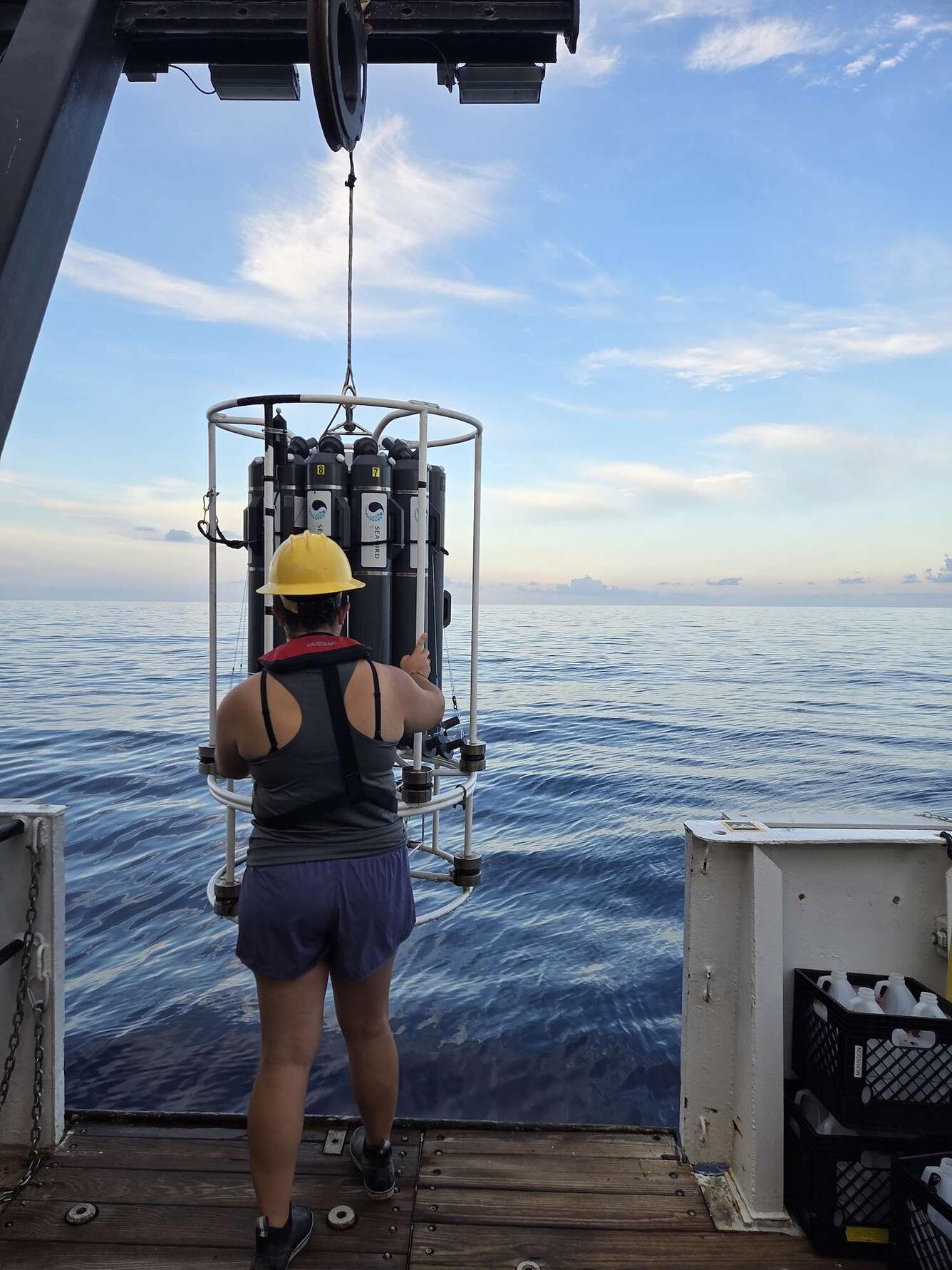 Woman in grey shirt and purple shorts with a yellow hardhat standing at the edge of the boat with the ocean in the background