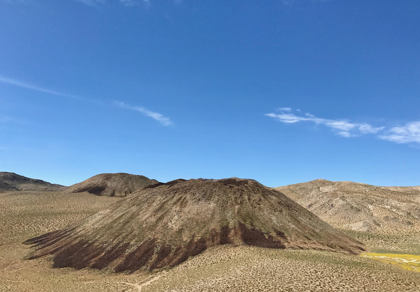 A photograph of several rocky volcanic hills shaped like upside-down flans. The hills and the landscape around them are dotted with sagebrush and dry grasses, revealing the underlying desert soil and rock. A bright blue sky overhead is streaked with clouds. USGS photo by Seth Burgess