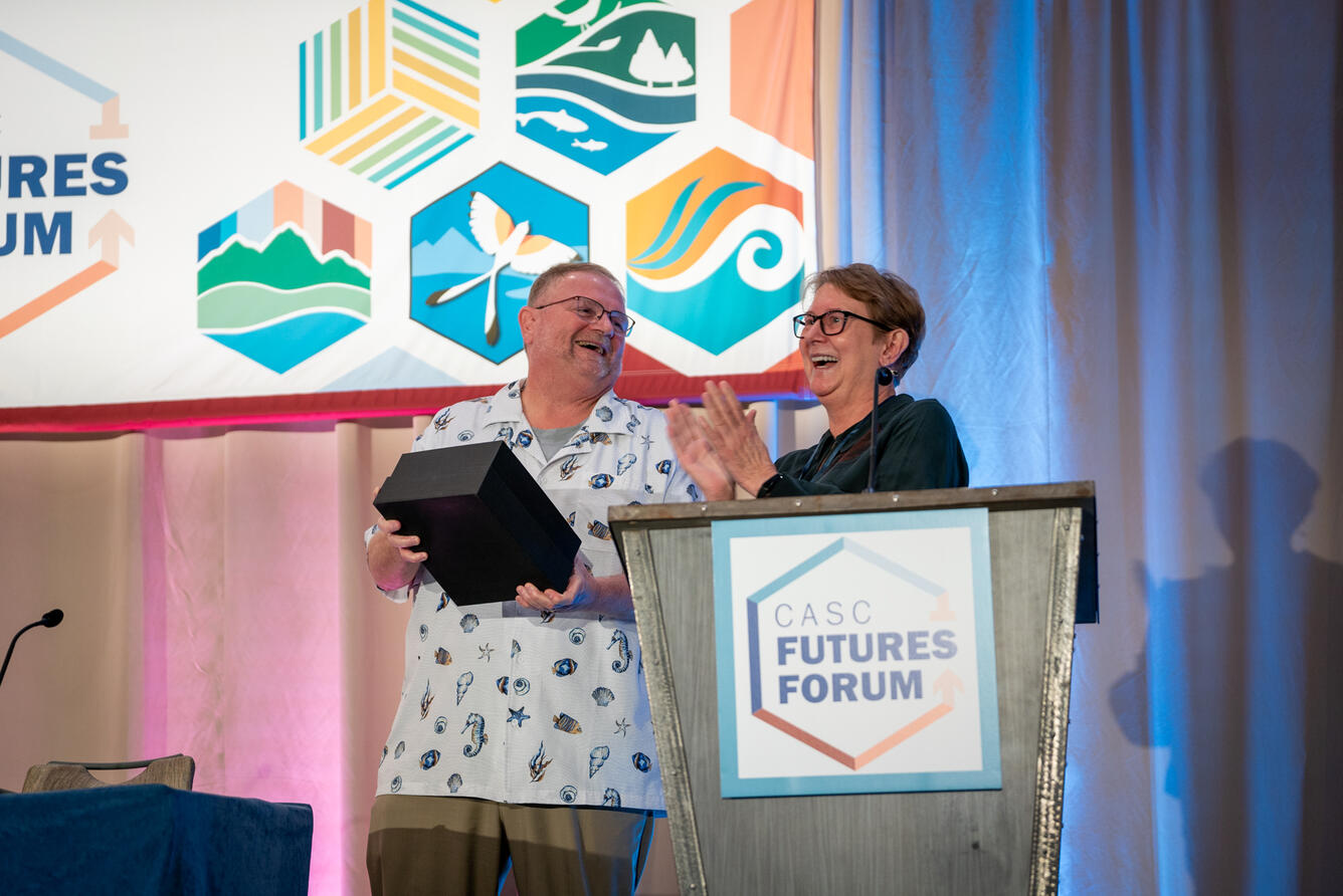 A man holding a plaque smiles at a woman behind a podium.