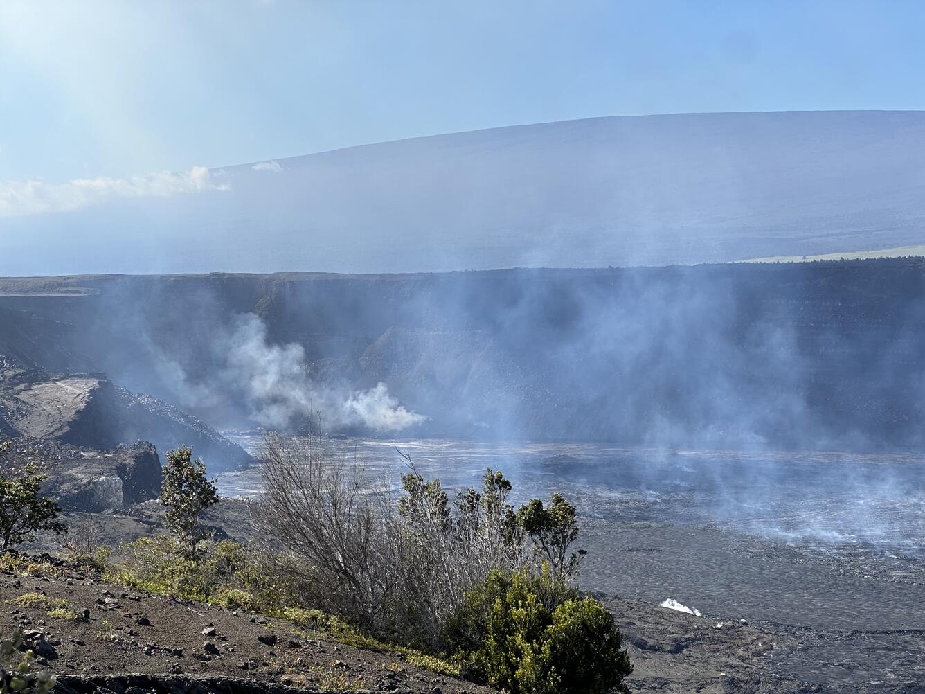 Hazy view of a crater wall with some vegetation in the foreground and a shield-shaped mountain in the background