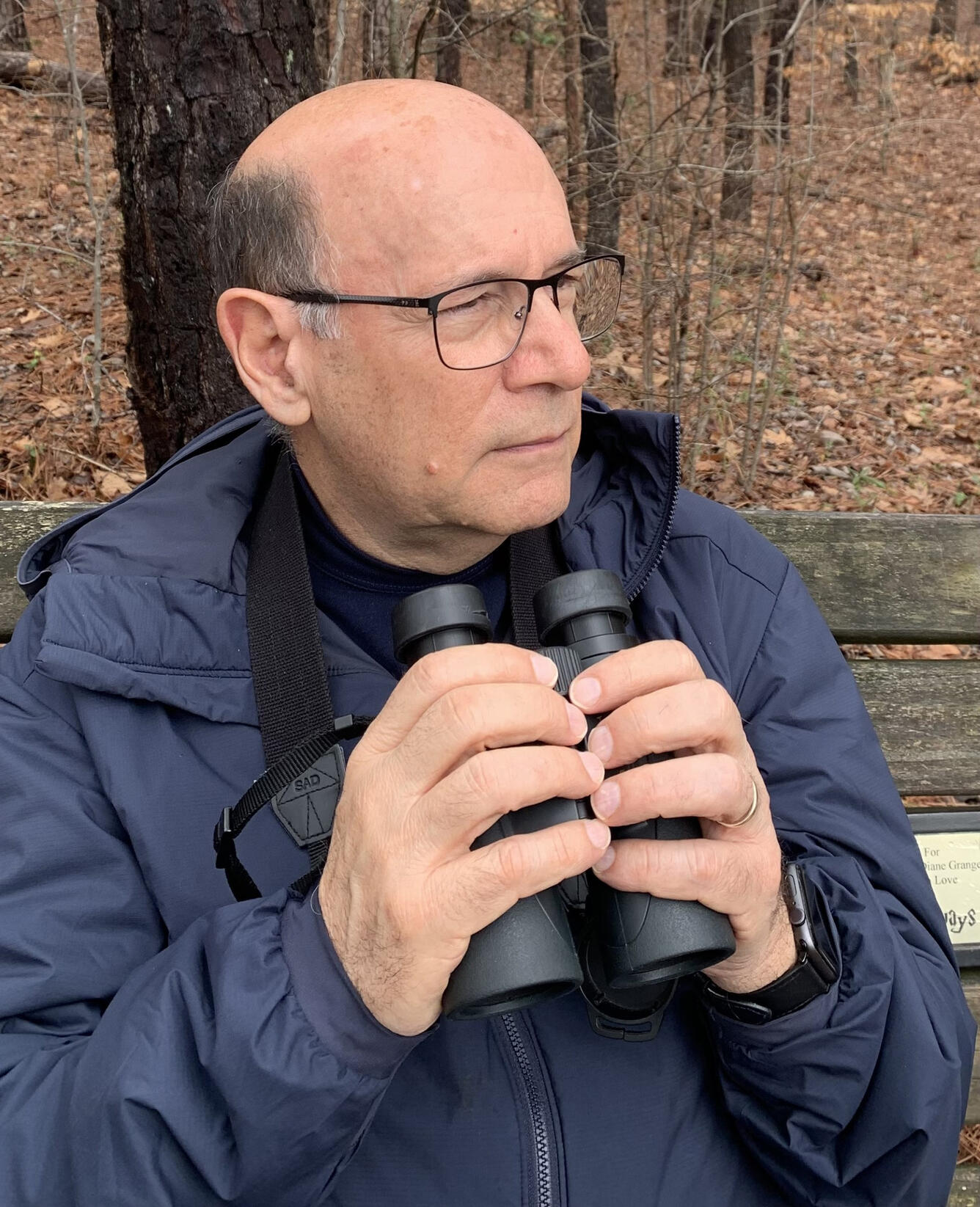 man wearing glasses and a blue jacket is standing in the woods holding binoculars