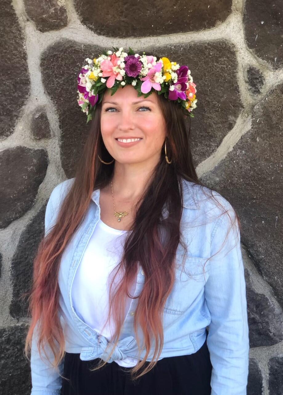 Person with long dark hair with a blue shirt in front of a stone wall wearing a flower crown lei