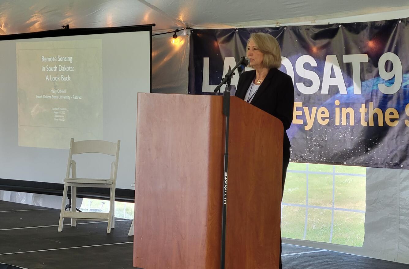 A woman speaking at a podium with a banner behind her and a screen beside her