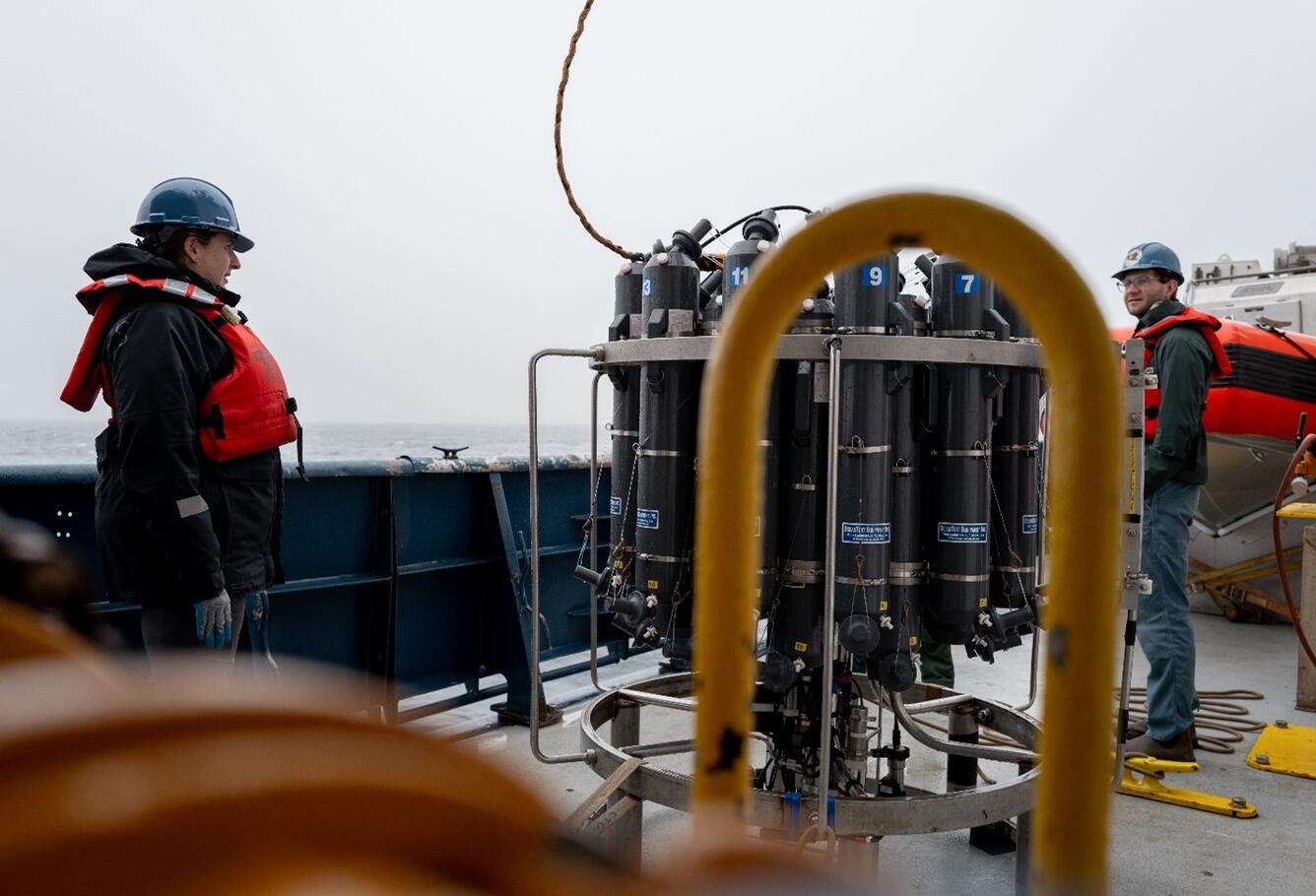 Two people in hard hats and life jackets stand on the deck of a research vessel next to the CTD