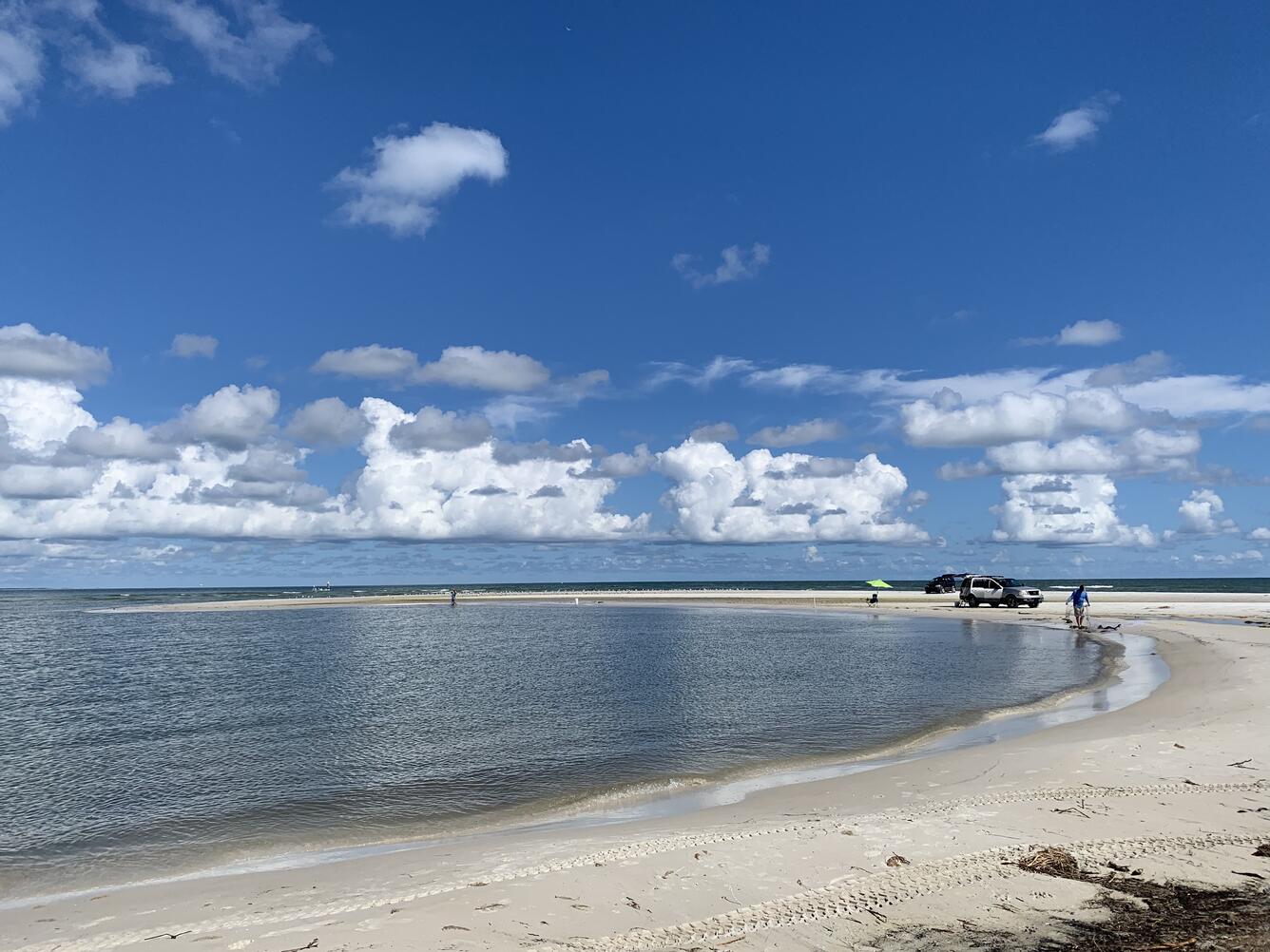 People and cars along a sandy beach, with water, blue sky and white clouds