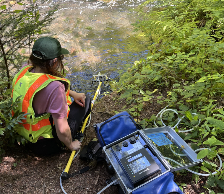lady inserts sampling bottles into stream water using rod from box-shaped device with tubes, battery pack, and pump for fill