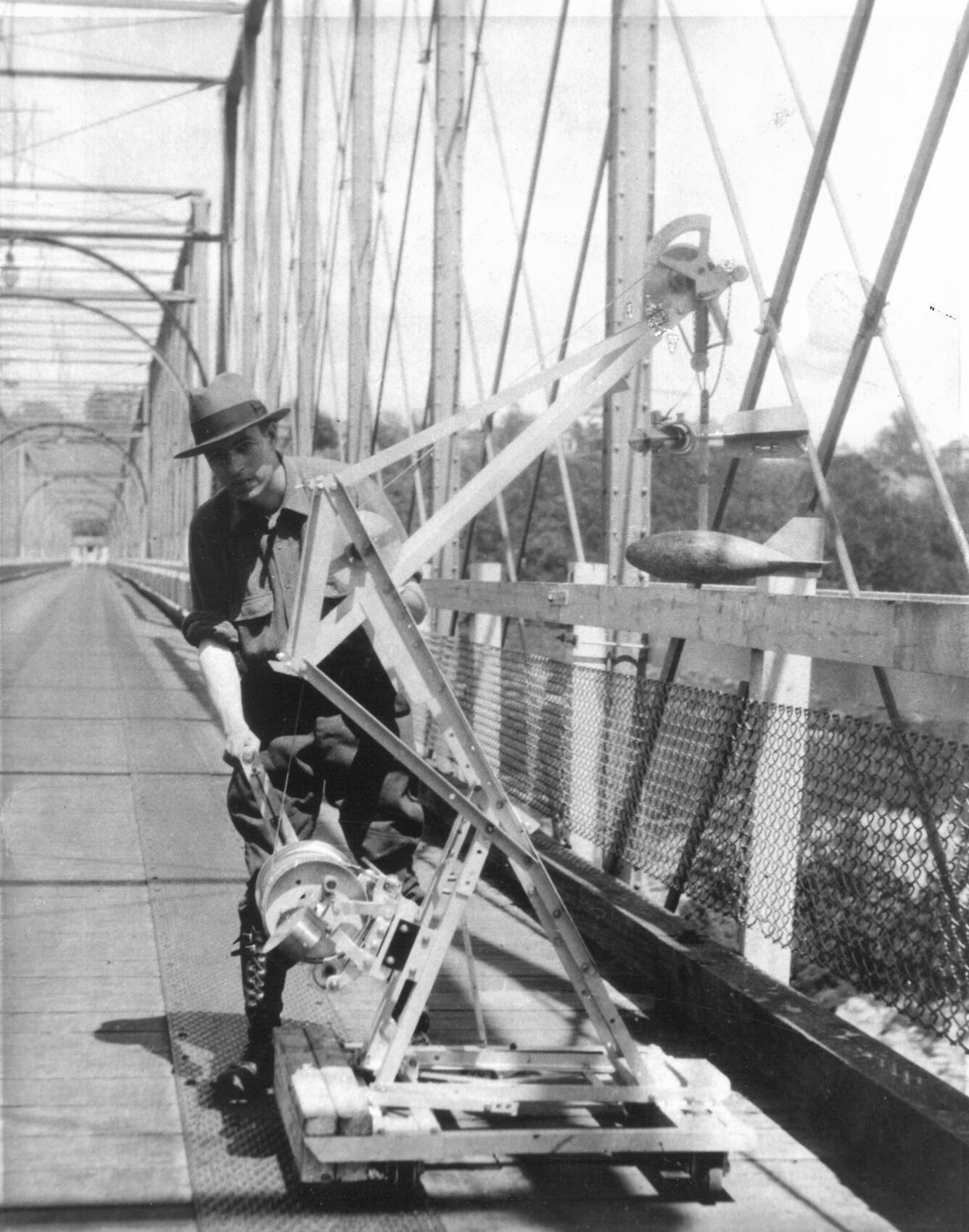 A black and white photo at the Chain Bridge along the Potomac River showing a technician posing next to a boom crane.