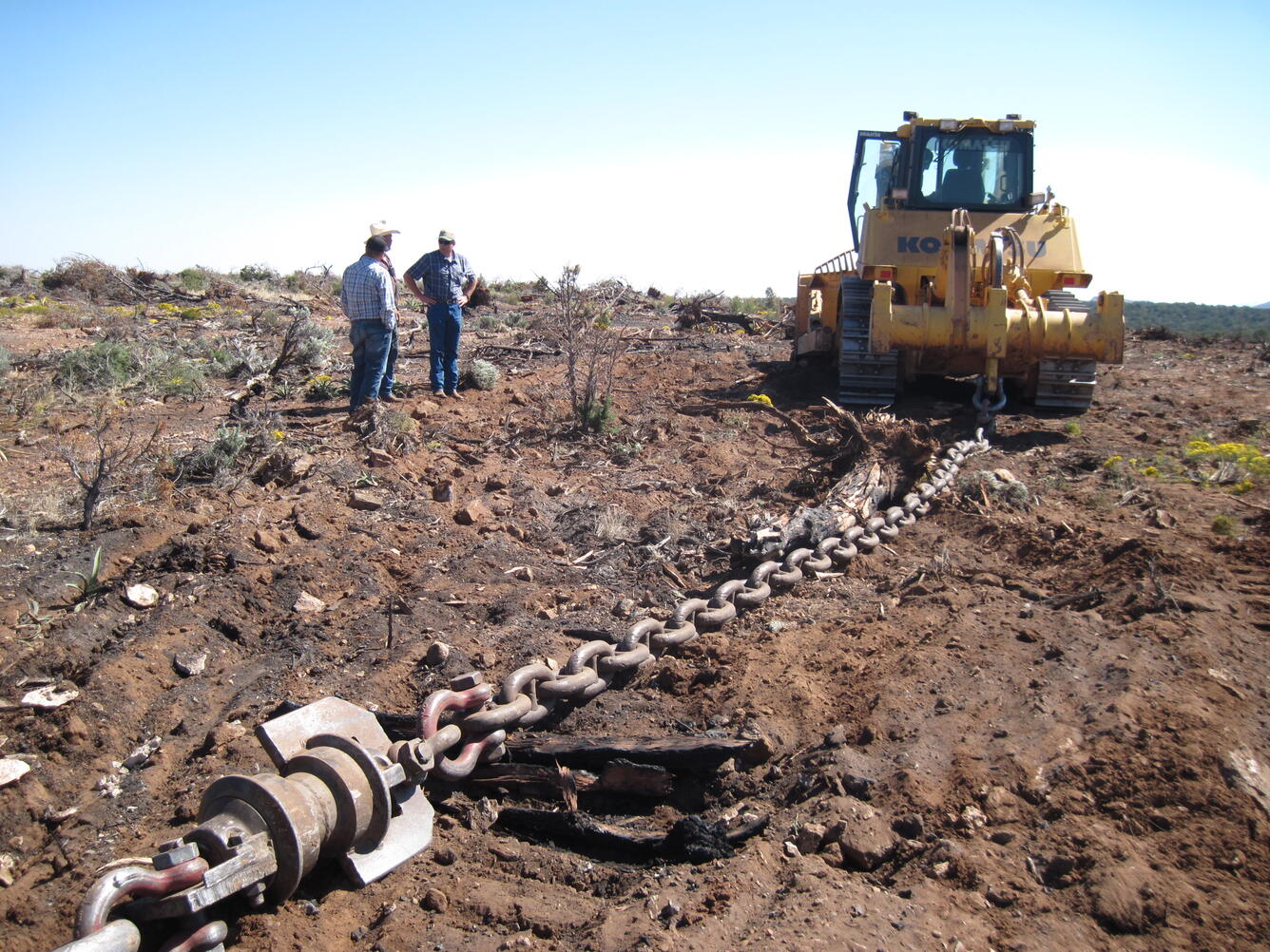 a tractor pulls a large train over muddy grounds. Three men stand nearby in conversation