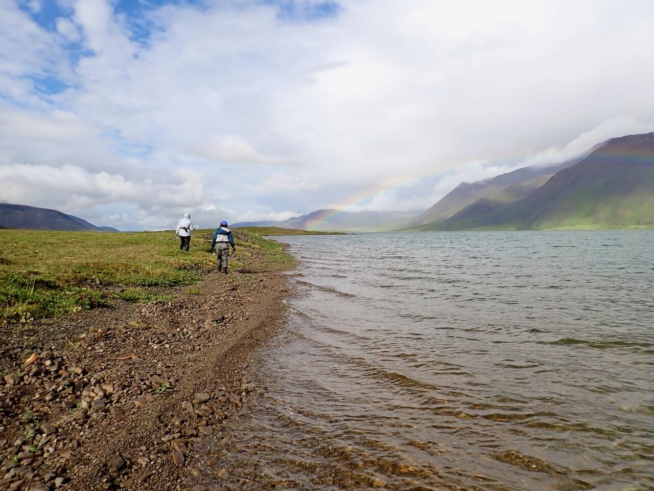 Two females on the grassy shoreline next to lake in rain gear and boots. Rainbow, blue sky with clouds and green mountains.  