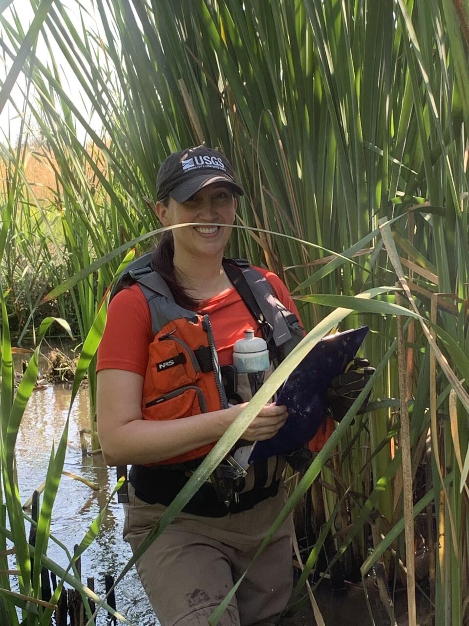 hydrologist stands in tall grassy vegetation in Chicken Creek holding a a clip board for note taking.