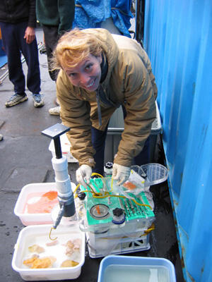 A person in a brown jacket leans over coral samples and smiles at the camera