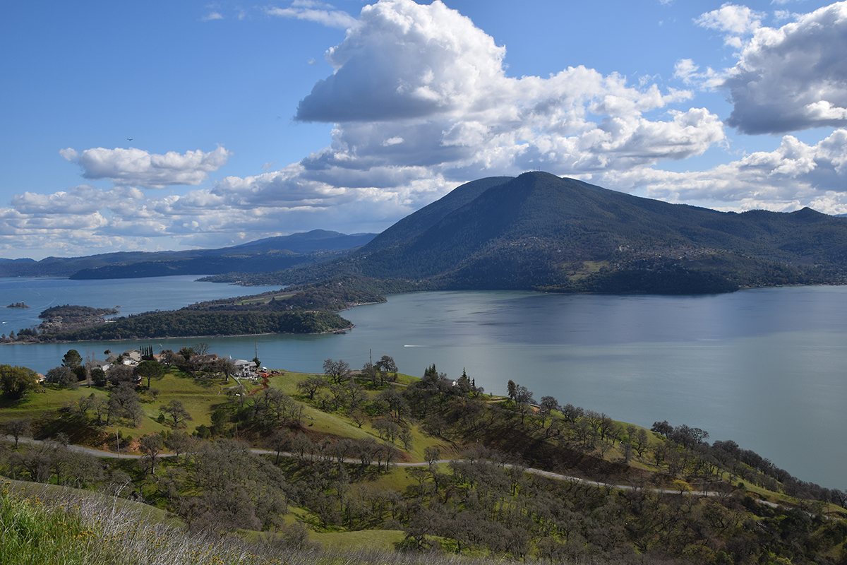 An expansive view from the top of a tall hill shows a large lake bordered by a large forested mountain with several peaks.