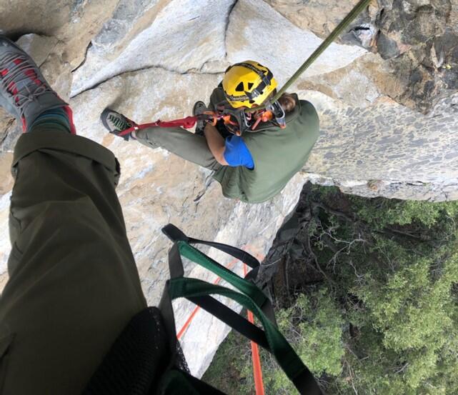 A rock climber on a rocky cliff looks down at another climber and the vegetation below