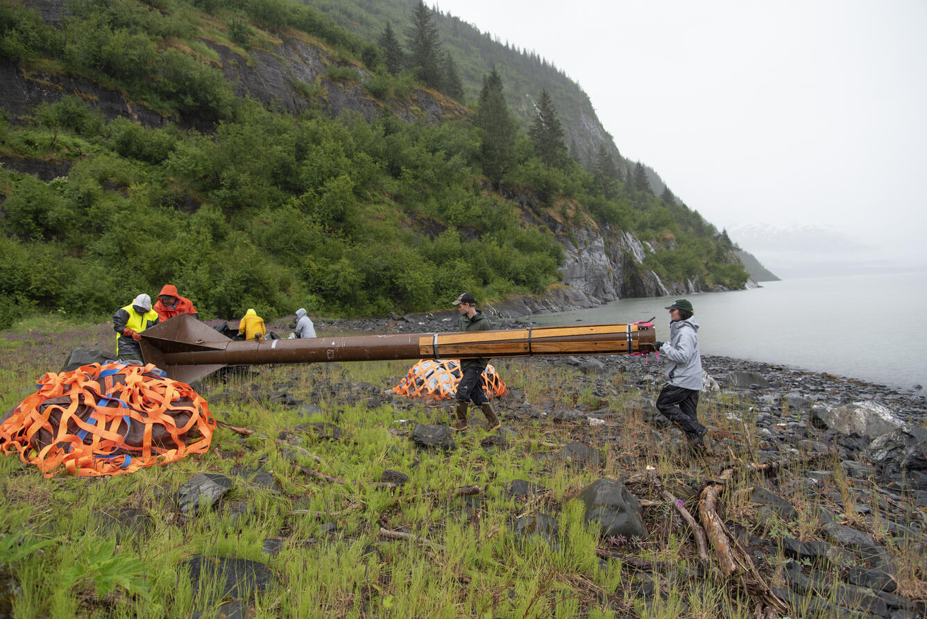 people installing large piece of equipment on shoreline of water body with ridge in background