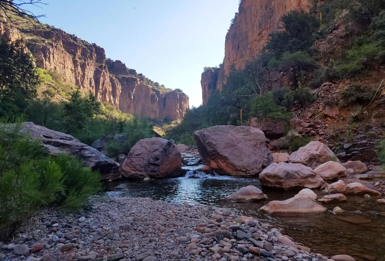 A creek with boulders and vegetation on the banks flows through a high desert canyon