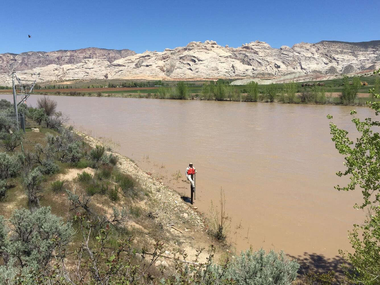 Hydrotech checks Crest Stage gage on edge of a wide Green River with green farmlands and white formations on opposite shore
