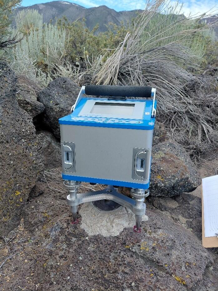 Box-shaped electronic instrument with a digital screen set up on rocky terrain surrounded by dry vegetation and mountains in the background.