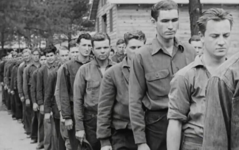 a historical image showing a line of young men in the Civilian Conservation Corps with a large wooden cabin and woods in the background