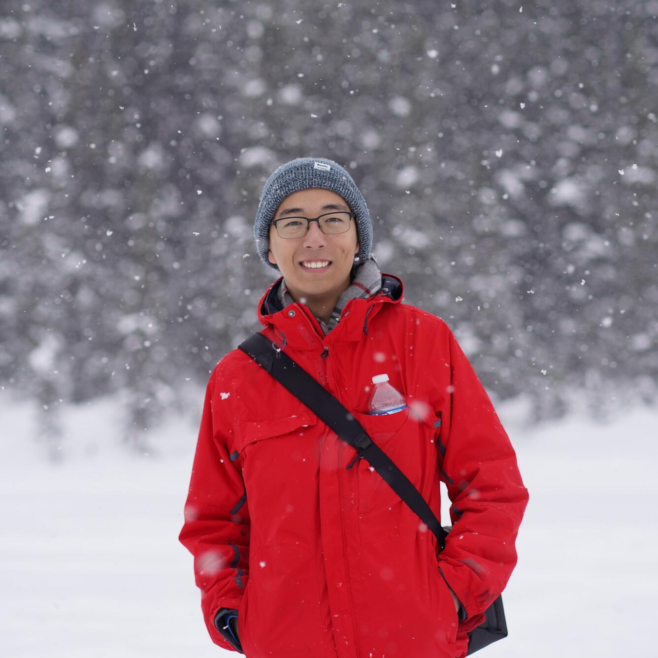 Zehao Xue stands in front of a snowy forest wearing a grey beanie and red jacket.