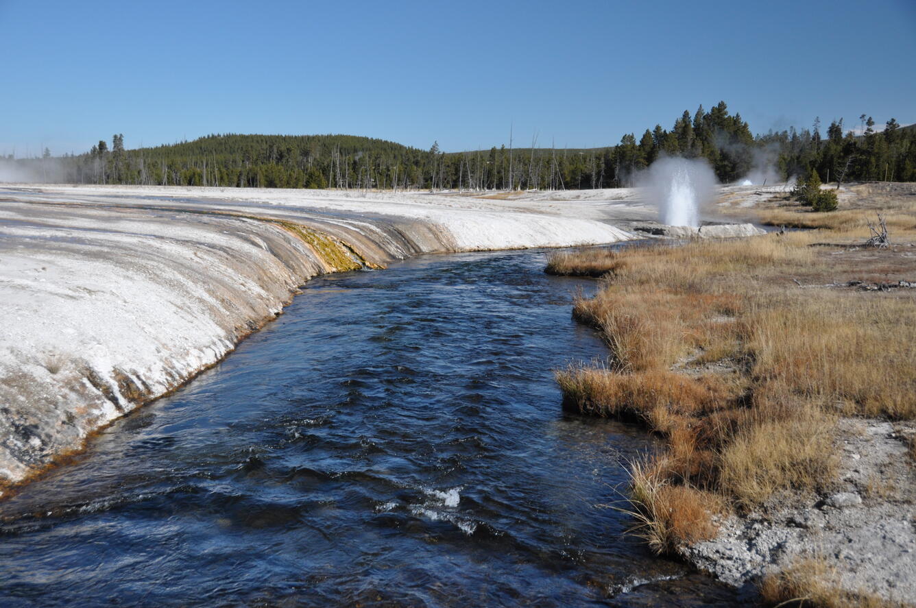 A stream winds pass a barren, white landscape on one bank and brown grass on the other; small geyser in the distance