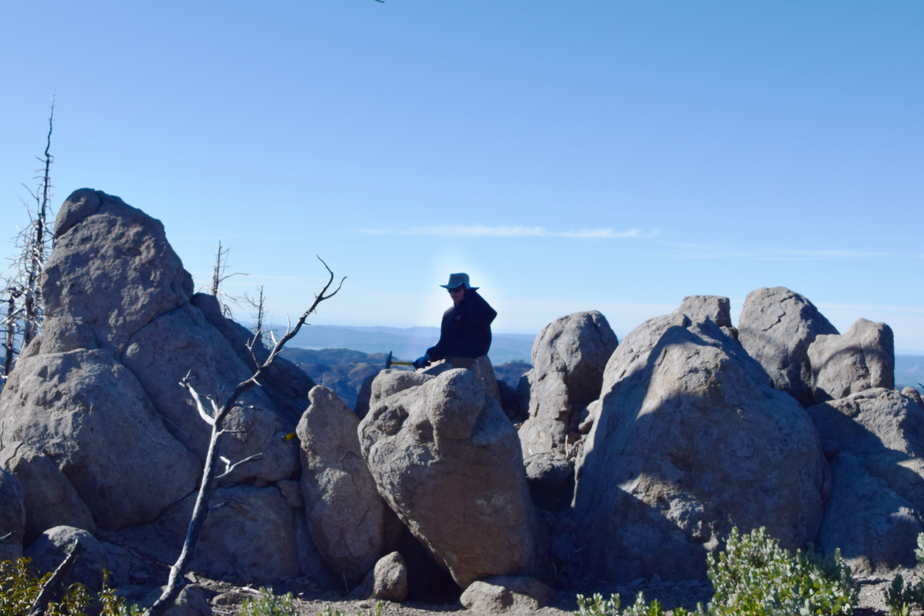 A scientist in a broad-brimmed hat sits on a large gray boulder of volcanic rock, surrounded by similar boulders. They are holding a rock hammer against the rock in preparation to break off a sample. In the distance, several hazy mountain ridges can be seen. 