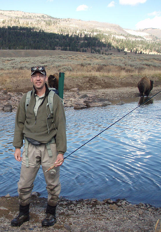 researcher holding a fly rod in yellowstone is standing near a bison along a river with mountains in the backdrop