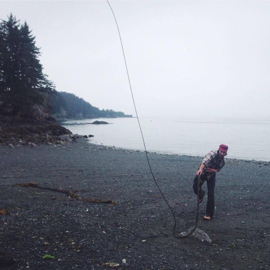 Man with fishing pole standing on gray beach casting fishing line.