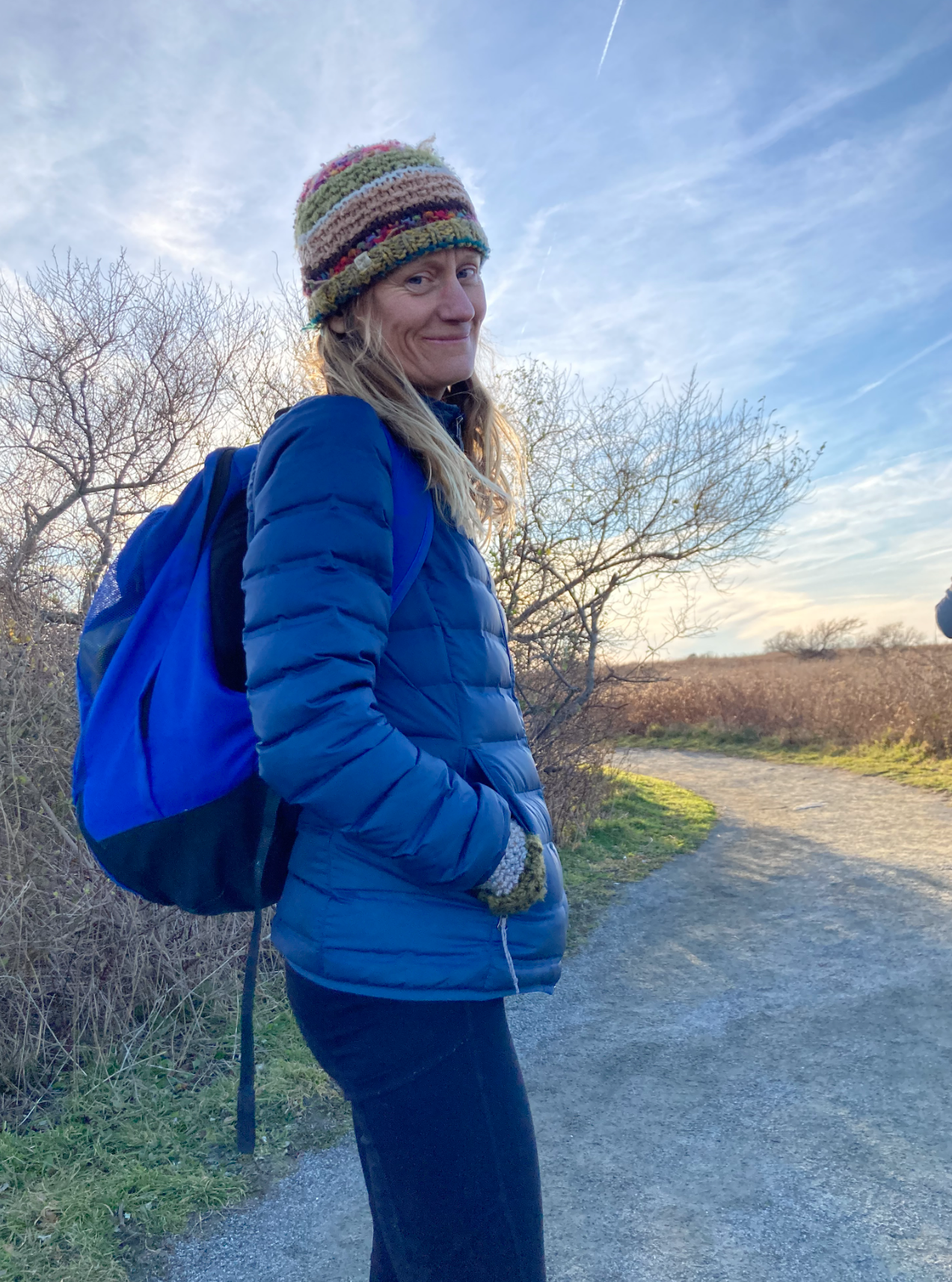 A woman in a hat and coat wearing a backpack stands on a tree-lined trail under a blue sky