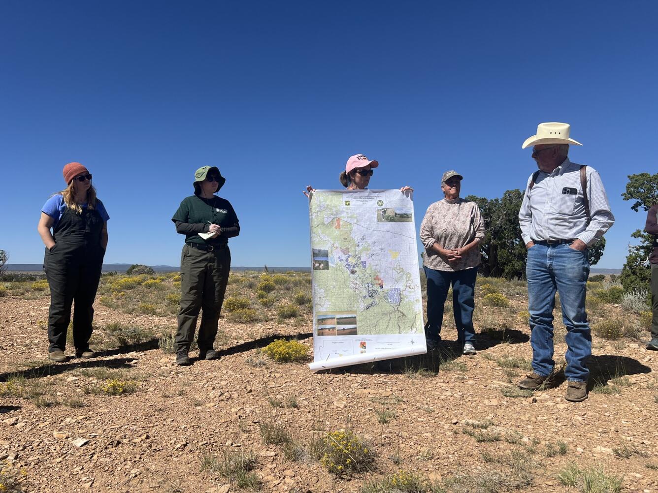 Five ranchers and researchers stand outdoors in a field looking at a large map, discussing land management