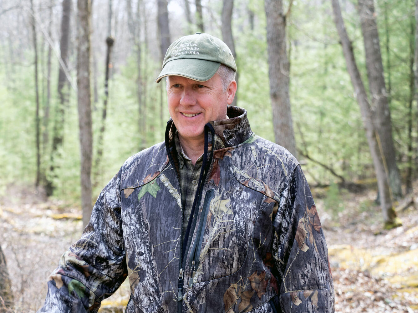 man wearing a camo jacket and green hat standing in the woods