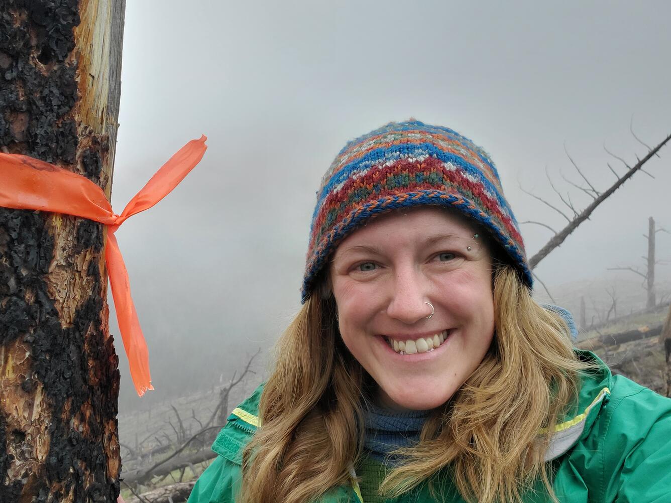 photo of person with long blonde hair in green jacket and multi-colored beanie, tree and clouds behind them