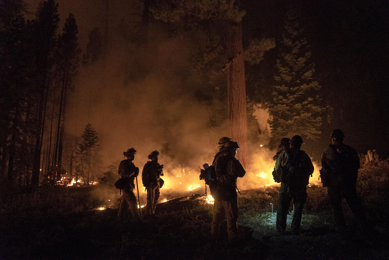 Flames and smoke are seen in a nighttime scene involving a handful of firefighters in silhouette
