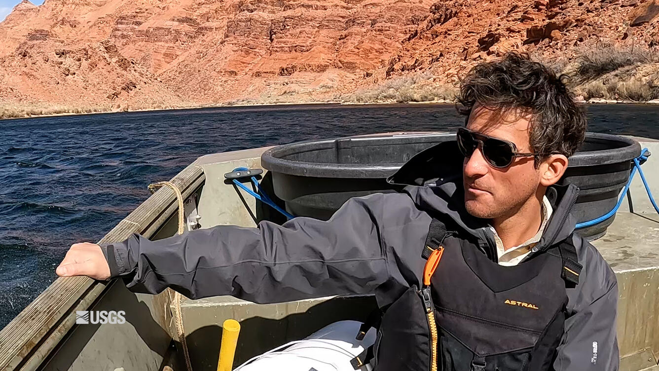 A man with curly hair and sunglasses grips the side of a boat as it drives along choppy waves through a red-rock canyon.