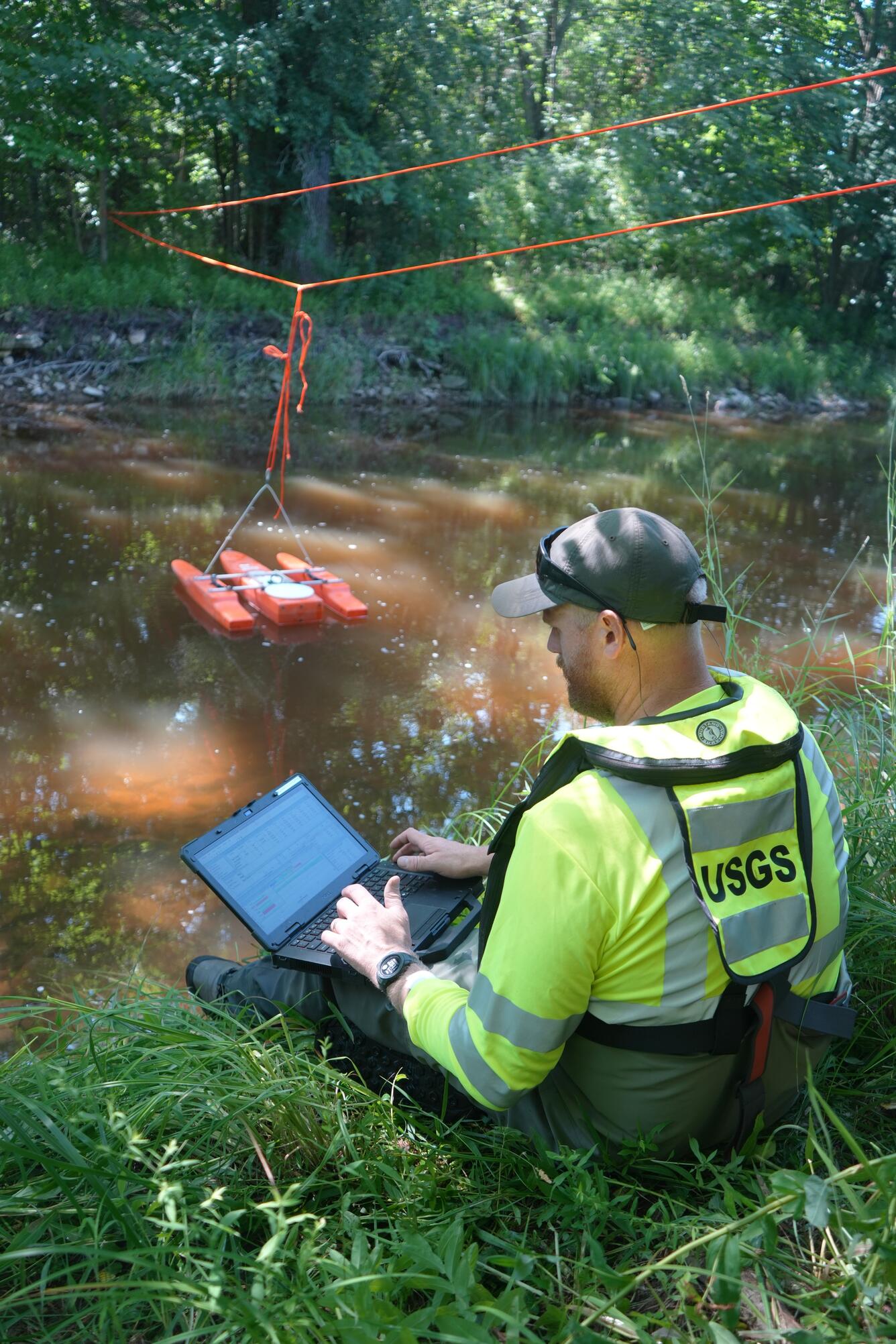 Hydrologic Technician sitting next to stream monitors streamflow data transmitted from an acoustic Doppler current profiler