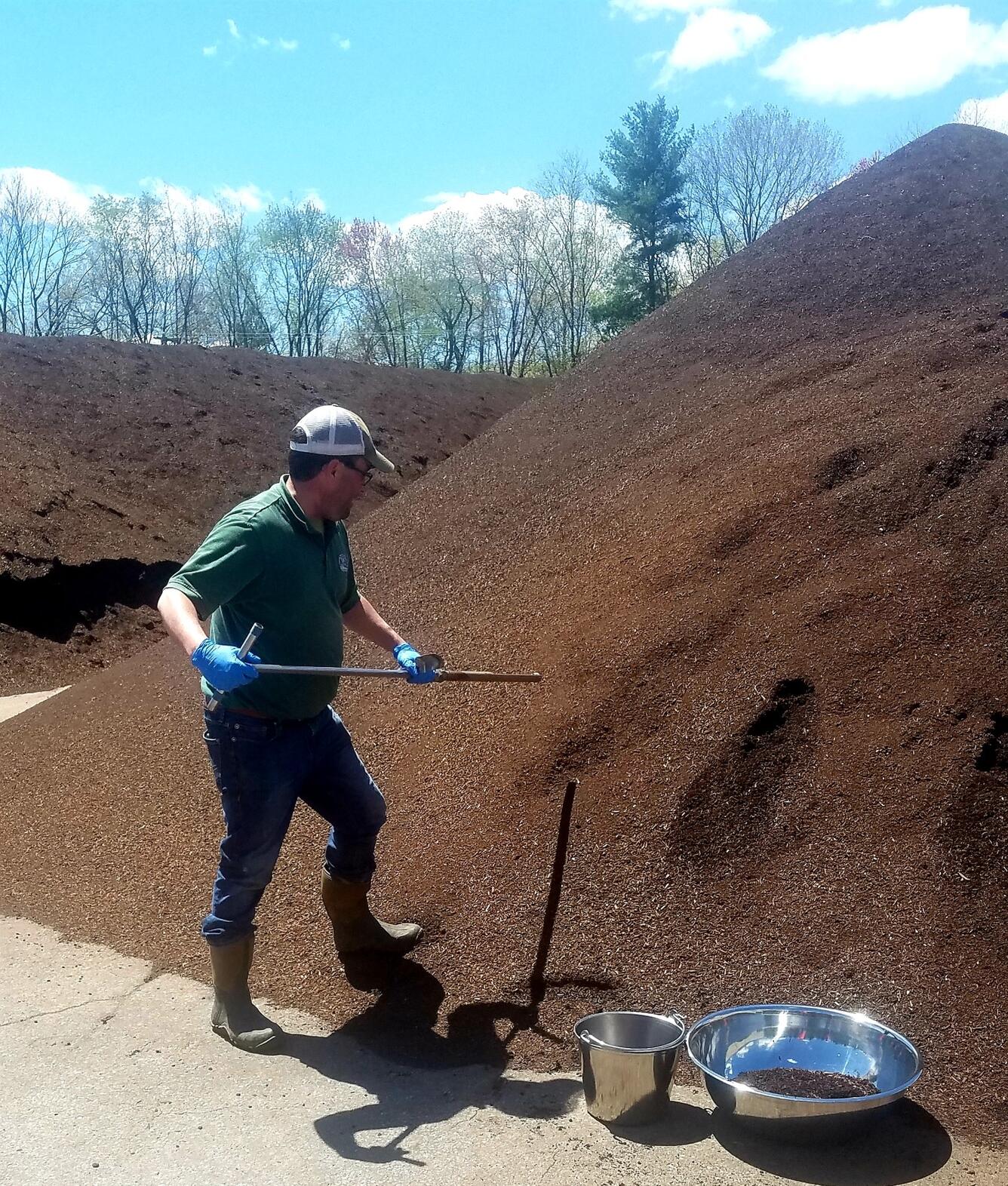 A man in green shirt and white baseball cap digs with shovel into a towering pile of dark brown dirt looking material