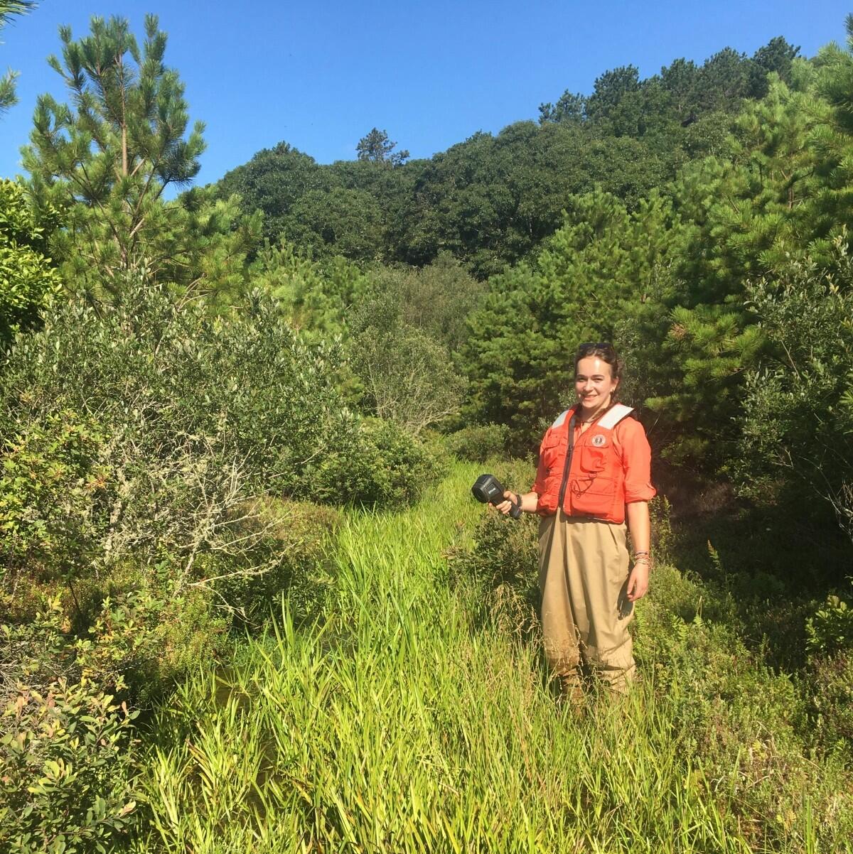 A lady in waders and orange life vest stands in a grassy bog surrounded by trees on a blue sky day