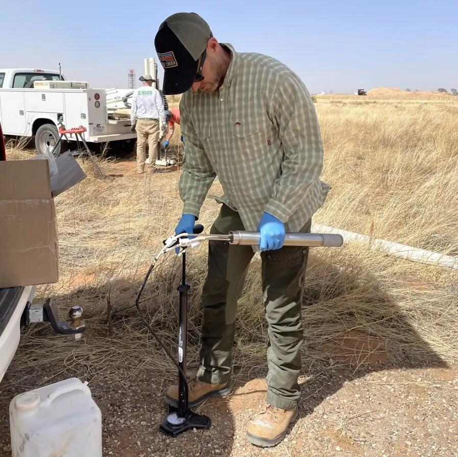 Male in baseball gap, glasses and latex gloved hands standing next to truck in a dry grassy field prepares an instrument