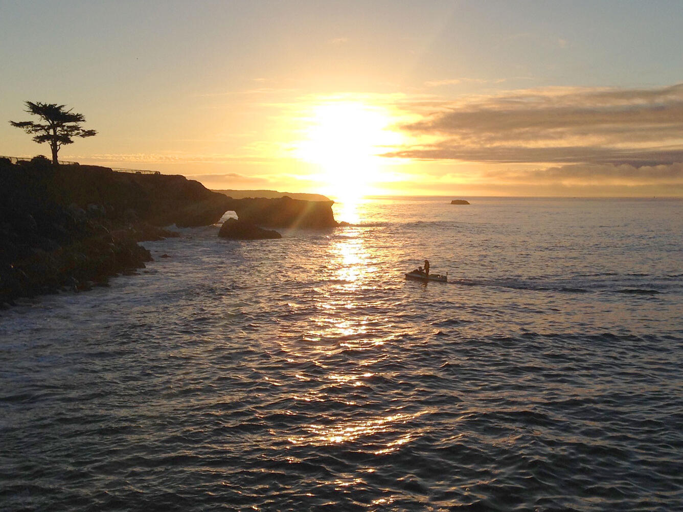 Sunset along a rocky coastline with a person riding a jet ski in the water.