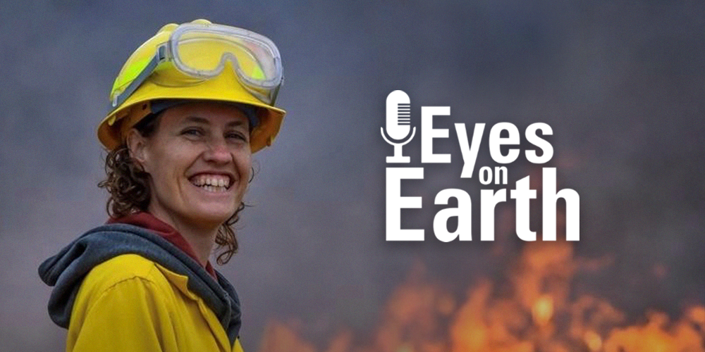 A woman smiles while dressed in a yellow suit with a fire burning in the background