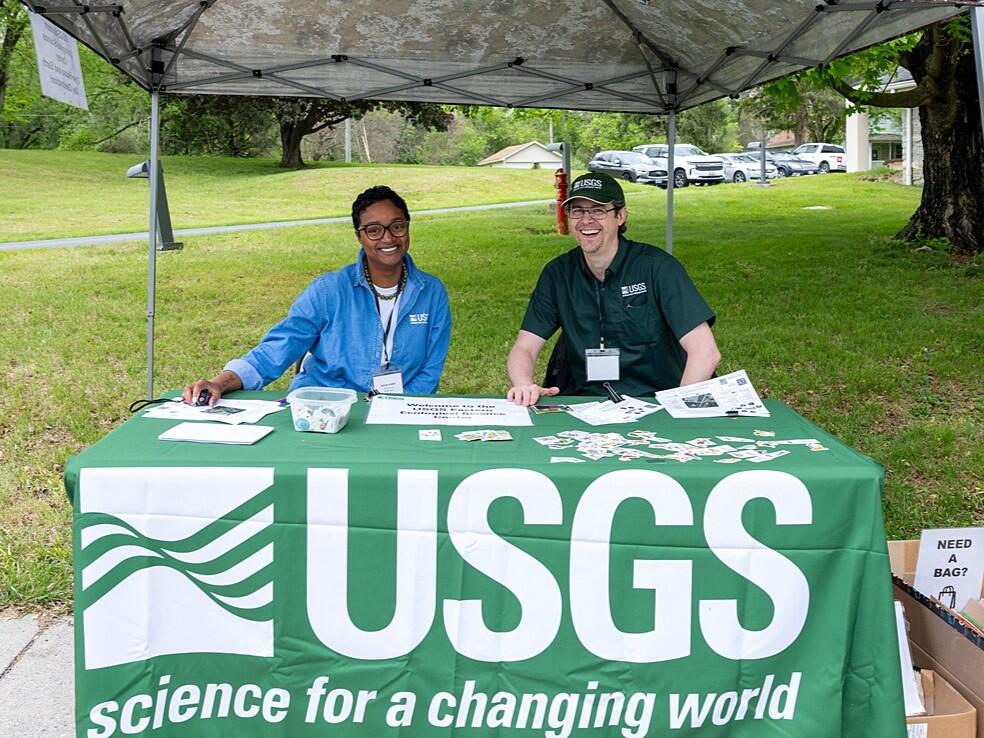 Two USGS employees smile while sitting behind a USGS tablecloth covered table under a pop up tent
