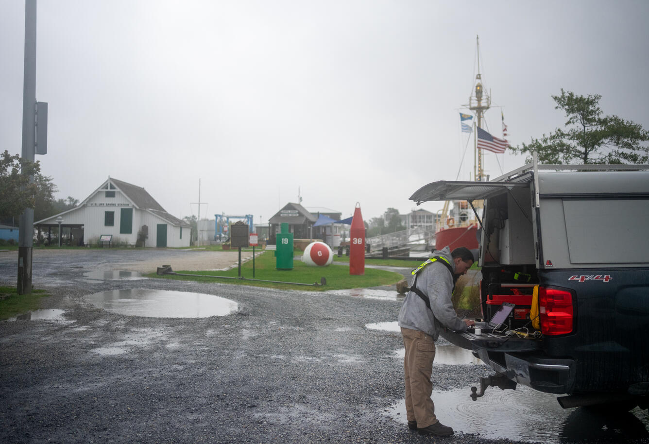 In a puddle ridden gravel parking lot, Chris Lewis takes refuge from the rain in the back of an SUV as he works on a laptop.