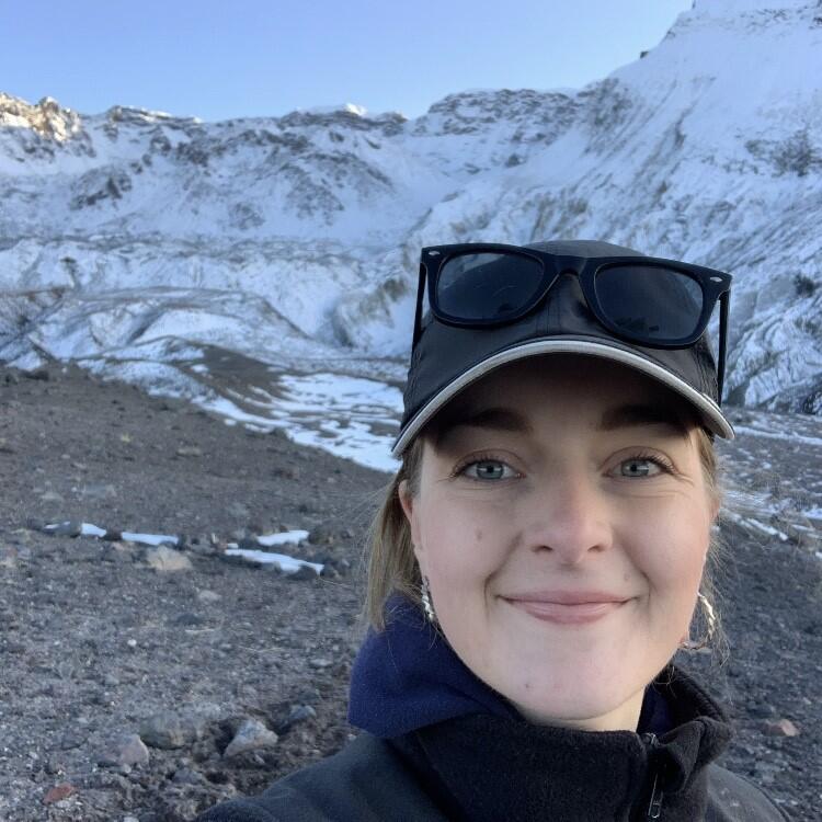 Woman standing in front of snowy mountain with cap and sunglasses on her head.